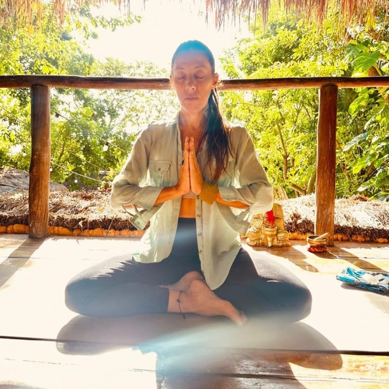Ximena Colombo en postura de meditación, con las manos en namasté, en una palapa rodeada de vegetación